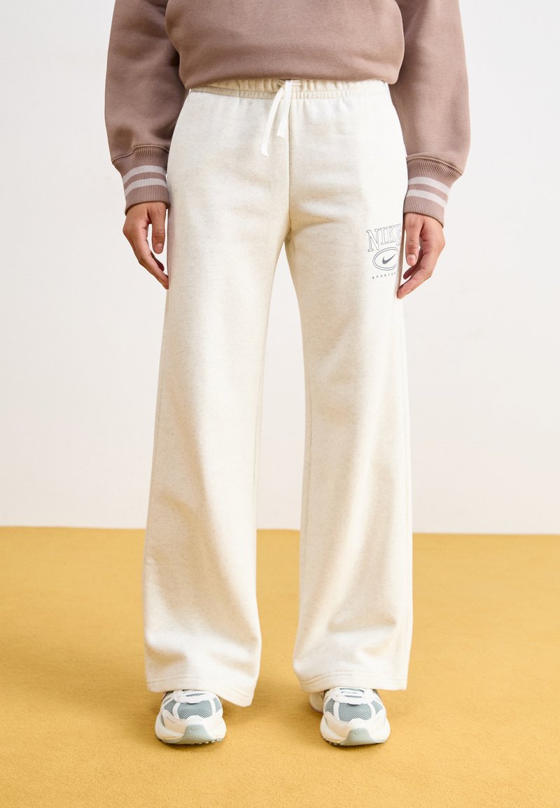 Fehér plüss melegítőnadrág húzózsinórral és nyomtatott Nike logóval. Szürke és fehér sportcipőkkel párosítva. Lapos, széles lábú dizájn.