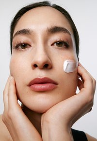 Zbliżenie twarzy o ciepłym odcieniu skóry, gładkiej teksturze, z nałożonym lekkim kremem na jednym policzku i delikatnie dotykającymi twarz dłońmi.