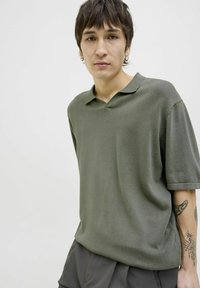 Polo tričko s krátkým rukávem v olivově zelené barvě s výstřihem do V a hladkou texturou. Model má na sobě šedé šortky a ukazuje tetování na paži.