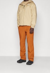 Man wearing beige North Face zip-up hooded jacket, burnt orange pants, and black hiking shoes standing against a white background.