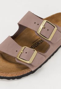 Sandalo in suede marrone con due cinghie regolabili. Presenta fibbie dorate e un footbed in sughero con suola in gomma nera.