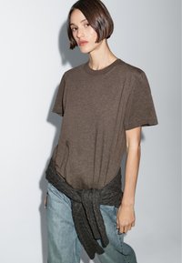 T-shirt in cotone marrone con maniche corte, vestibilità rilassata e collo rotondo. Un maglione a maglia grigio scuro è annodato in vita. Jeans in denim chiaro.