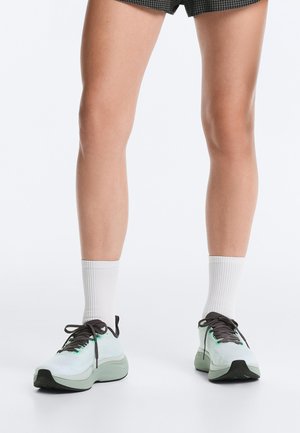 Picioare care poartă pantaloni scurți negri în carouri, șosete albe până la gleznă și adidași atletici gri deschis cu șireturi negre pe fundal alb.