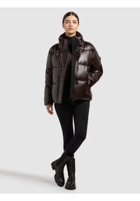 Brun dunjacka med hög krage, glansig yta och knäppning framtill. Bärs över en svart topp och svarta leggings, kombinerat med svarta stövlar.