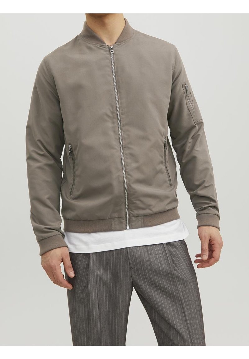 Homme portant une veste bomber grise zippée avec poches zippées, un t-shirt blanc et un pantalon gris à fines rayures sur fond uni.
