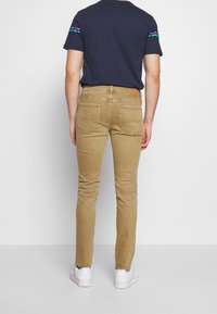 Man som bär smala beige jeans, en marinblå t-shirt med lila och turkosa ränder på ärmarna, och vita sneakers, som står framför en enfärgad vit bakgrund.