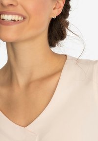 Guldtonad studörring med en texturerad cirkulär design och en klar ädelsten i mitten, mot en ljusrosa tygbakgrund.