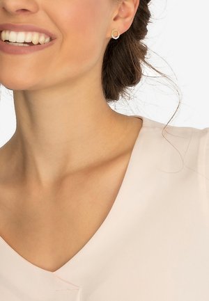 Guld-tone stud ørering med et tekstureret cirkulært design og en klar ædelsten i midten, sat mod en lyserød stofbaggrund.