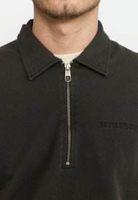 Zwarte ritsblouse met een geribbelde textuur, voorzien van een kraag, contrasterende rits en geborduurd "REVOLUTION" logo op de borst.