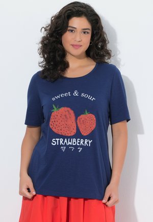 Kvinde med krøllet mørkt hår iført en marineblå skjorte med to store jordbær og teksten "sødt & surt JORDbær nyd hver dag."