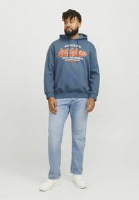 Marineblå hættetrøje med hvid og orange logo tekst, parret med lyseblå jeans og hvide sneakers. Hætten er foret og har ribkantede ærmer.