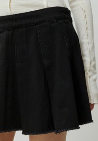 Schwarzer Jeansrock mit gefälteltem Design und fransigem Saum. Der Bund ist leicht gerafft und legt eine strukturierte Oberfläche offen.