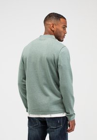 Graugrüner Pullover mit Kragen, strukturiertem Stoff und langen Ärmeln. Getragen über einem weißen Shirt, kombiniert mit dunkelblauen Jeans.