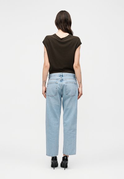 Γυναίκα με μακριά καστανά μαλλιά, που φοράει μαύρο αμάνικο τοπ, ανοιχτό μπλε τζιν με ίσια γραμμή και μαύρα ψηλοτάκουνα παπούτσια, γυρισμένη ανάποδα.