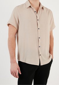 Camisa beige de manga corta hecha de tela texturizada, con cierre frontal de botones azul oscuro y corte relajado.