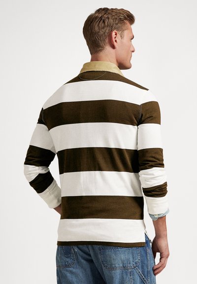 Vīrietis no aizmugures, tērpies brūnganā un baltā platu svītru garroku polo krekliņā un zilās džinsās ar uzlocītām piedurknēm.