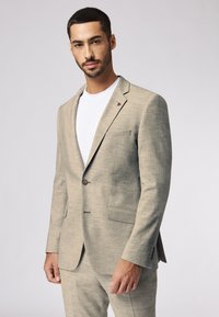 Hellgrauer Blazer aus strukturiertem Stoff, mit einem einzigen Knopfverschluss und zwei vorderen Taschen. Mit einem kleinen roten Emblem am Revers.