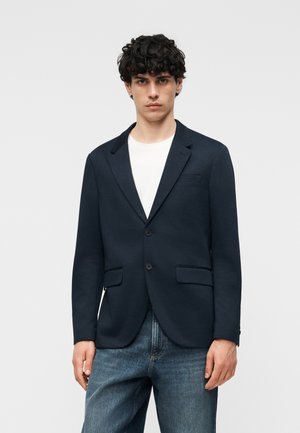 Nuori mies kiharatukka, yllään tummansininen bleiseri valkoisen paidan ja sinisten farkkujen päällä, seisoo yksinkertaista vaaleaa taustaa vasten.