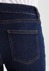 Dunkelblaue Jeans mit orangefarbener Ziernaht, einer Gesäßtasche mit quadratischem Design und einer glatten Denim-Textur. Gürtelschlaufen am Bund sichtbar.