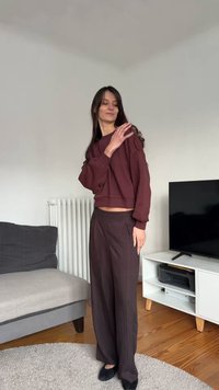 Bordowy krótki sweter z balonowymi rękawami, w połączeniu z szerokimi brązowymi spodniami. Czarne buty. Proste, wewnętrzne pomieszczenie z jasnymi meblami.