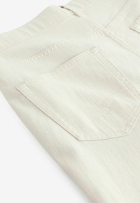 Ljusbeige denimtyg med en synlig bakficka. Texturen ser slät ut, och det finns subtila sömdetaljer längs kanterna.