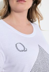 Camiseta blanca con un cuello redondo, que presenta letras en negro con puntos que forman la 'Q' y un patrón de puntos negros formando una figura triangular en el frente.
