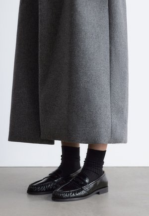 Personne portant des chaussettes noires et des mocassins noirs brillants avec une jupe grise longueur cheville, debout sur un sol lisse.