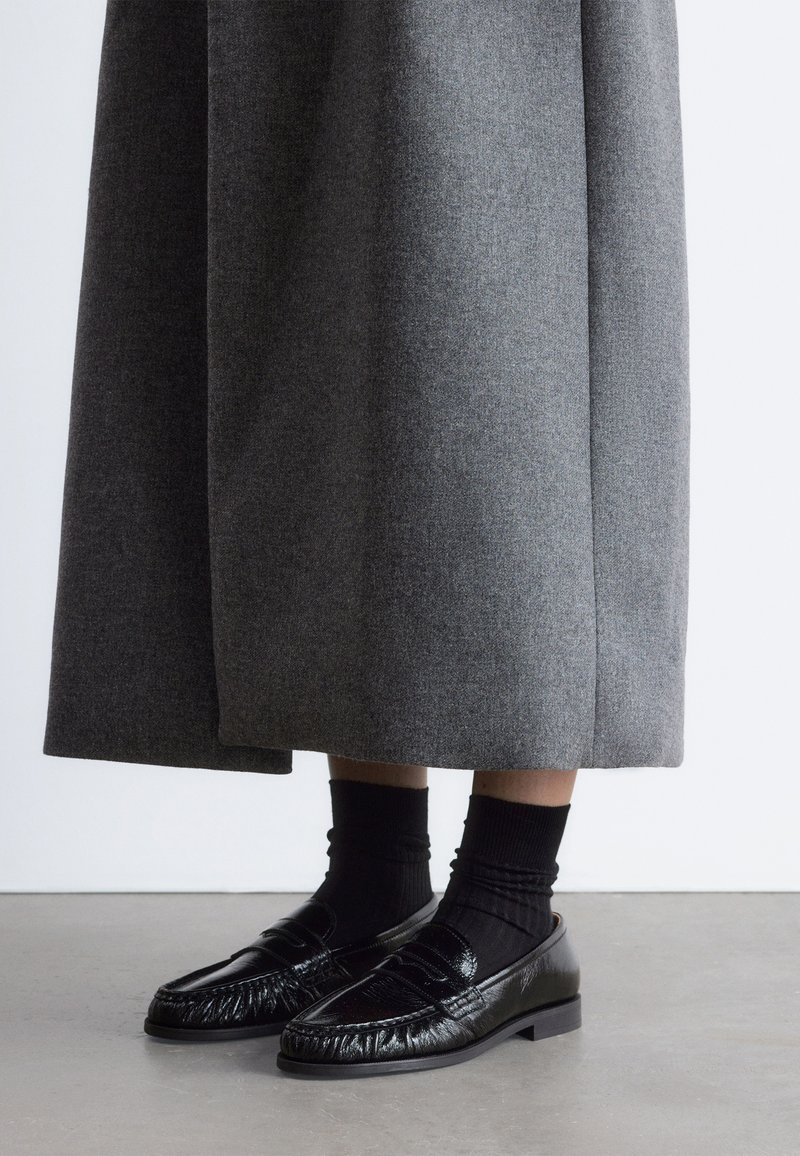 Personne portant des chaussettes noires et des mocassins noirs brillants avec une jupe grise longueur cheville, debout sur un sol lisse.