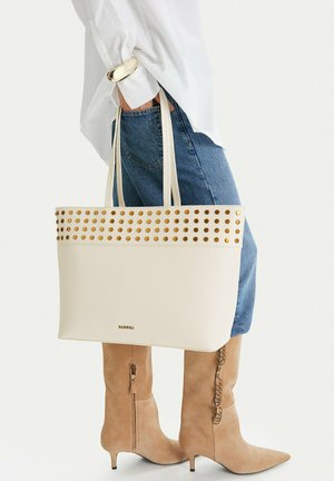 Persona vistiendo jeans azules, botas beige hasta la rodilla, camisa blanca, sosteniendo un gran bolso tote crema con tachuelas doradas y logo en el frente.