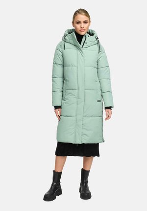 Woman wearing a long light green puffer coat with hood, black ribbed skirt, and black chunky boots, standing on a plain white background.