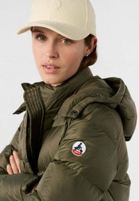 Giacca bomber verde oliva con colletto alto e cappuccio; presenta una toppa con il logo sulla manica. Indossata con un cappello beige chiaro in corda.