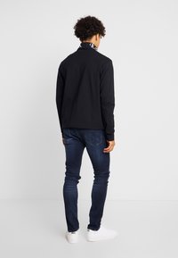 Camisa negra de manga larga con cuello con marca, combinada con jeans ajustados azul oscuro y zapatillas blancas, mostrando un estilo casual moderno.