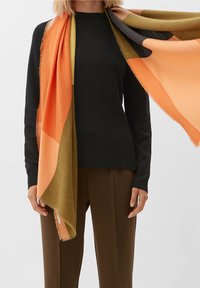 Écharpe en laine aux couleurs orange, olive et gris foncé avec des bords à franges, drapée sur un haut noir à manches longues, assortie à un pantalon marron.