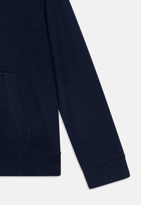 Haut en hoodie texturé bleu marine avec une poche kangourou, des poignets et un ourlet côtelés, arborant un motif en tissu tissé. Vue en gros plan de la manche et de la poche.