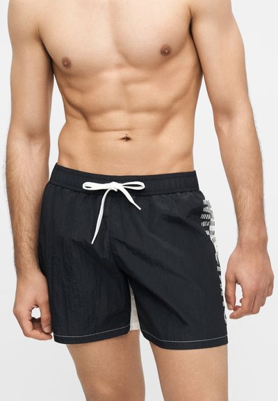 Ανδρικός κορμός με μυώδη κατασκευή που φοράει μαύρο μαγιό με λευκό κορδόνι και λεπτομέρεια λευκής ρίγας στο πλάι, σε απλό φόντο.