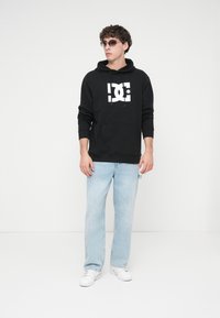 Black cotton hoodie with a front pocket and white graphic logo. Worn with light blue denim jeans and white sneakers. Model also in sunglasses.