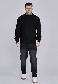 Must triibuline kudumist kampsun, ümmarguse kaelusega, kombineeritud tumedate teksade ja valgete tennistega. Käte ja kaela piirkonnas on nähtavad tätoveeringud. Lihtne ja puhas disain.