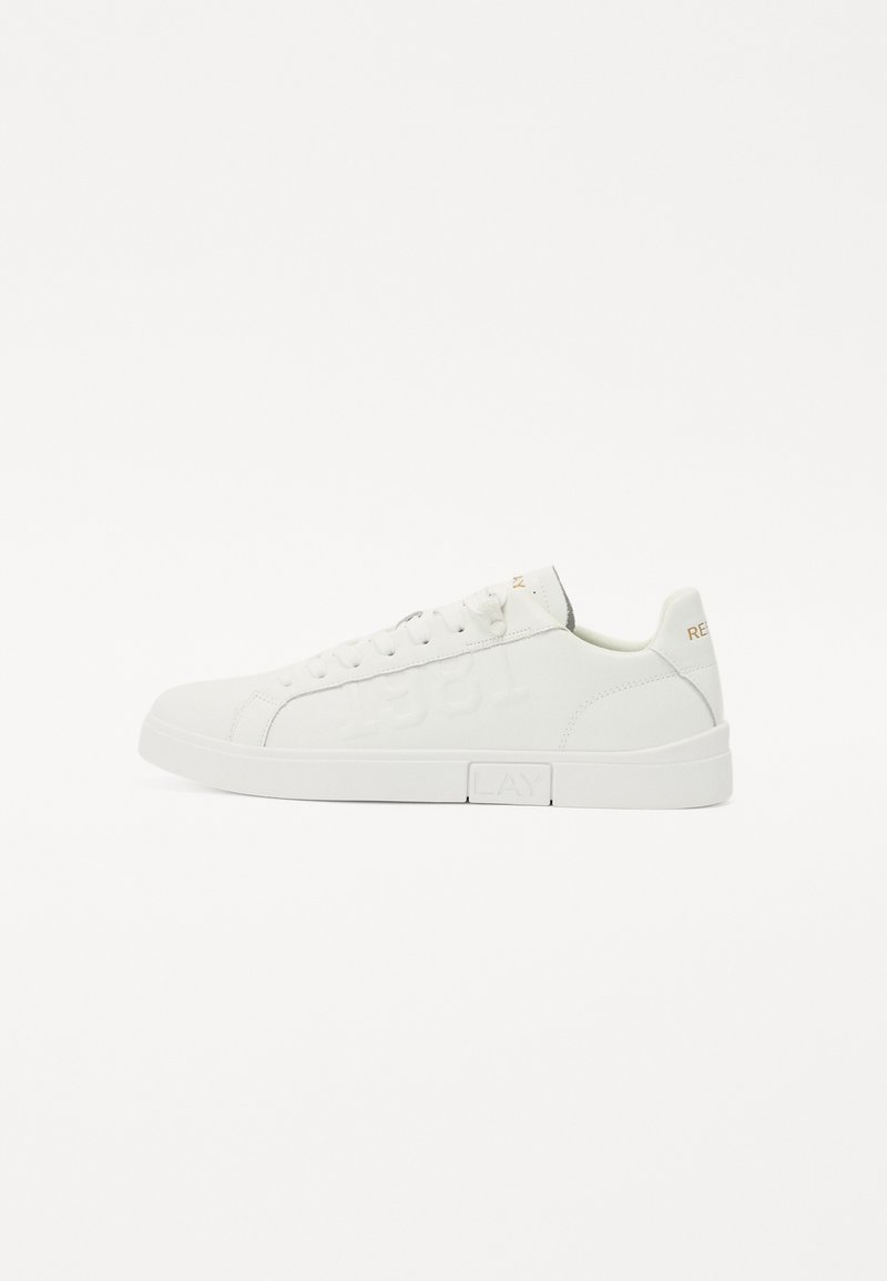 Fehér sneaker texturált bőr felsőrésszel, kerek orral és lapos gumitalppal. Hímzett számokkal és logóval rendelkezik, kontrasztos színekben.