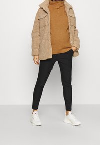 Veste en polaire beige, pull marron côtelé, et pantalon noir à rayures. Baskets blanches avec des touches de rose. Accent sur la texture et le contraste des couleurs.