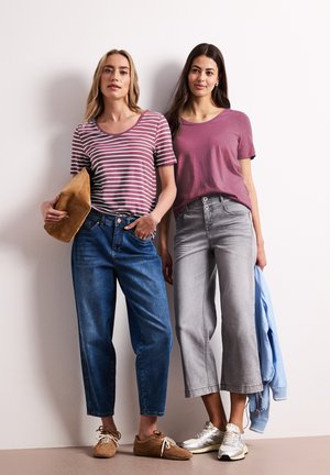 To kvinder står op af en væg; den ene i en stribet lilla skjorte med blå jeans og holder en brun taske, den anden i en lilla skjorte med grå korte jeans og holder en jakke.