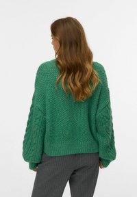 Pull en tricot vert avec une texture épaisse et des détails en tricot torsadé sur les manches. Il a une coupe décontractée et un ourlet côtelé. Porté avec un pantalon gris.
