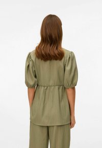 Blusa verde chiaro, tessuto testurizzato, con maniche a sbuffo, vita elastica e design fluido. Il retro presenta una finitura liscia e arricciata.