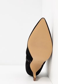 Chaussure à talon stiletto en daim noir à bout pointu, inclinée pour montrer la semelle beige lisse et la pointe étroite du talon sur fond blanc.