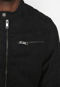 Chaqueta de ante negro con cuello alzado, cierre frontal de cremallera y bolsillo en el pecho con herrajes de tono plateado y costura detallada.