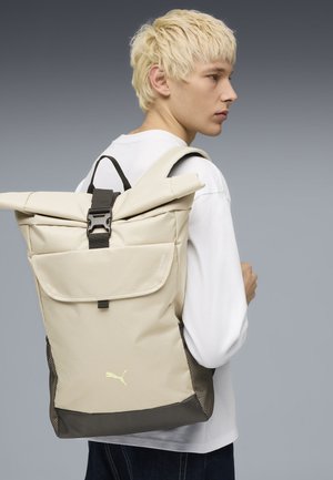 Ung person med kort blondt hår iført hvit skjorte og beige roll-top ryggsekk med svart spenne mot grå bakgrunn.