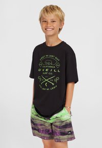 Camiseta negra con diseño gráfico verde que presenta un oso y texto; combinada con pantalones cortos de baño multicolores en patrones de olas verdes y moradas.
