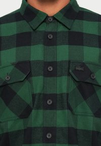 Camicia a quadri verde e nera in tessuto, con chiusura a bottoni frontale, due tasche sul petto con patta e un dettaglio del logo.