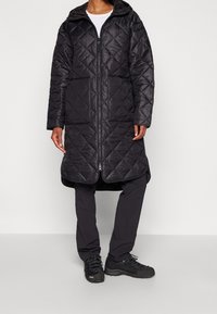 Abrigo acolchado negro con capucha, que cuenta con un cierre frontal y mangas largas. Llevado con pantalones oscuros y calzado negro resistente.