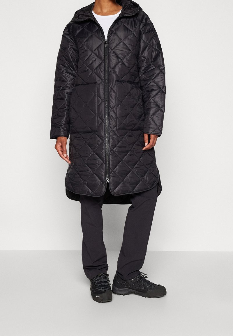 Abrigo acolchado negro con capucha, que cuenta con un cierre frontal y mangas largas. Llevado con pantalones oscuros y calzado negro resistente.