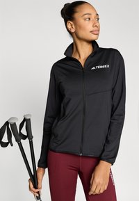 Fekete cipzáras dzseki magas gallérral, „TERREX” logóval. Bordó leggingsszel viselve, túrabotokkal kiegészítve.